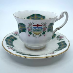 May include: A white teacup and saucer set with a green, yellow, and red plaid pattern and a crest featuring a mountain range and a flag. The set is decorated with pink flowers.