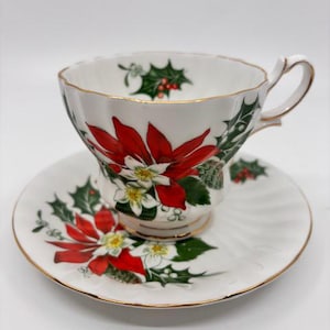 May include: A white teacup and saucer set featuring a Christmas poinsettia design. The cup and saucer are decorated with red poinsettias, green holly leaves, and white flowers.