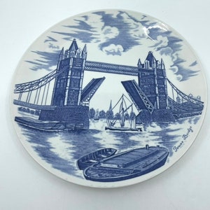May include: A blue and white ceramic plate featuring a depiction of Tower Bridge in London. The bridge is drawn with the drawbridge raised, and there are boats in the water below. The text "Tower Bridge" is written in blue at the bottom right of the plate.