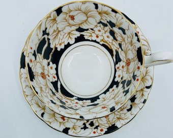 May include: A black and white teacup and saucer with a floral pattern. The cup and saucer have a gold trim and feature a large white flower with red accents.
