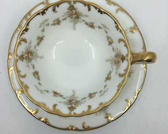 May include: White teacup and saucer set with gold trim and floral accents. The cup and saucer have a scalloped edge and a gold handle. The floral design features small, muted orange and green flowers. The set is likely vintage or antique.