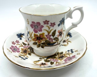 Può includere: Un set da tè bianco con tazza e piattino con un motivo floreale in tonalità di rosa, giallo, blu e verde. La tazza e il piattino hanno un bordo dorato.