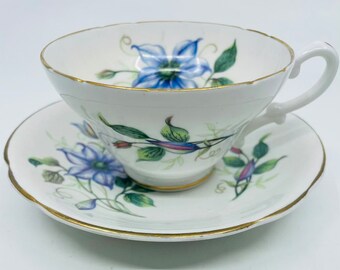 May include: White teacup and saucer set with a floral design. The teacup and saucer feature a blue flower pattern with green leaves and gold trim. The teacup has a curved handle and a scalloped edge.