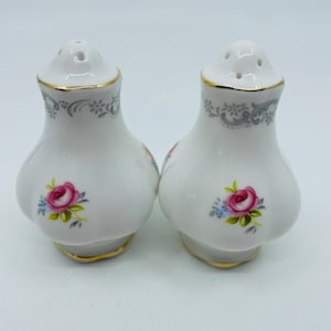 Könnte beinhalten: Ein Set aus zwei weißen Salz- und Pfefferstreuern aus Keramik mit floralem Design. Die Streuer haben einen goldenen Rand und zeigen rosa Rosen und blaue Blumen.