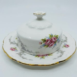 Puede incluir: Un mantequillero de porcelana blanca con un diseño floral. El mantequillero tiene una tapa con un pomo en la parte superior. El mantequillero está decorado con rosas rosas y azules y hojas grises. El mantequillero tiene un borde dorado.
