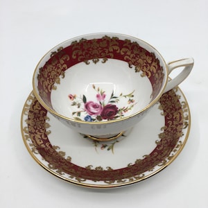 Può includere: Una tazza da tè e un piattino bianchi con un designo floreale rosso e dorato. La tazza presenta un motivo floreale con rose rosa e rosse. Il piattino ha un bordo dorato e un designo floreale rosso e dorato.