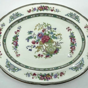 Può includere: Piatto ovale da portata con un motivo floreale. Il piatto in ceramica bianca presenta un bouquet centrale di fiori viola, rossi e blu. Un bordo verde e grigio circonda la composizione floreale e il bordo del piatto.