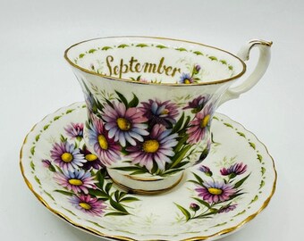 May include: A white teacup and saucer with a floral design. The teacup has the word "September" written on it in gold lettering. The flowers are pink, purple, and green.