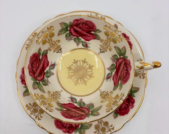 May include: A vintage teacup and saucer set with a floral design. The cup and saucer are white with a yellow center and feature red roses with green leaves. The edges of the cup and saucer are trimmed with gold.