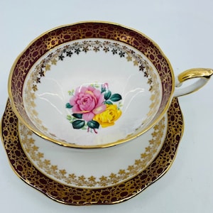 May include: A white teacup and saucer with a burgundy and gold rim. The cup and saucer feature a floral design with pink and yellow roses.