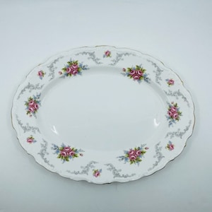 Könnte beinhalten: Eine weiße ovale Servierplatte mit einem zarten Blumenmuster. Das Muster zeigt rosa Rosen, blaue Blätter und ein graues, wirbelndes Design. Die Platte hat einen goldenen Rand.