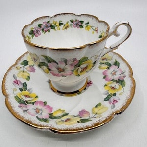 May include: A white teacup and saucer set with a floral pattern in shades of pink, yellow, and green. The set has a gold trim around the edges.