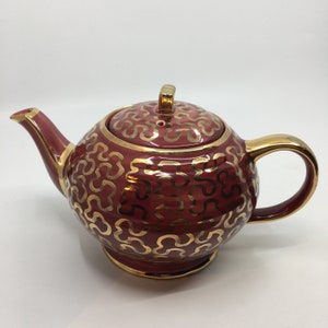 May include: A red teapot with a gold pattern of stylized clover shapes. The teapot has a lid with a gold knob.