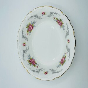 Könnte beinhalten: Eine weiße Keramikschüssel mit einem gewellten Rand und einem floralen Design. Die Schüssel hat einen goldenen Rand und zeigt rosa und graue Rosen mit grünen Blättern.