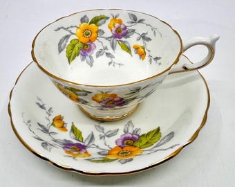May include: A white teacup and saucer set with a floral design. The cup and saucer are trimmed with gold and feature orange and purple flowers with green leaves. The floral pattern is set against a white background.