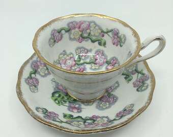 May include: A white teacup and saucer with a floral pattern in shades of pink, yellow, and green. The edges of the cup and saucer are trimmed with gold.