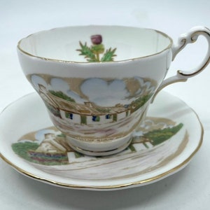 May include: A white teacup and saucer set with a gold trim. The teacup features a painted image of a cottage with a thatched roof and a green bush. The saucer has a similar image of a cottage and a green bush. The teacup and saucer are decorated with a purple thistle.