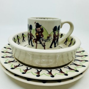 May include: A white ceramic children's dish set with a marching band design. The set includes a bowl, a plate, and a mug.