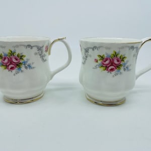 Könnte beinhalten: Zwei weiße Keramikbecher mit einem Blumendesign mit rosa Rosen und blauen Akzenten. Die Becher haben einen goldenen Rand und ein graues Blumenmuster.