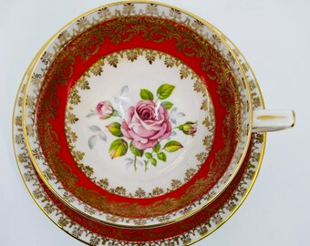 Pode incluir: Um conjunto vintage de chávena e pires com um design floral vermelho e dourado. A chávena apresenta uma rosa rosa com folhas verdes e uma borda dourada. O pires tem um design correspondente.