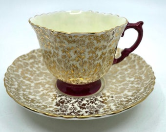Può includere: Una tazzina da tè e un piattino bianchi con un motivo floreale dorato e un bordo color granata. La tazza ha un bordo ondulato e un manico color granata.