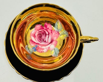 May include: A gold teacup and saucer set with a black rim and a pink rose design.