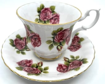 May include: A white teacup and saucer set with a pink rose floral pattern. The cup and saucer have a gold trim around the edges.