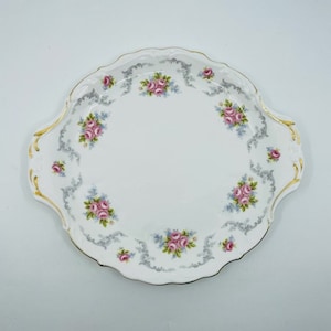 Könnte beinhalten: Ein weißer Keramik-Kuchenteller mit einem gewellten Rand und einem zarten Blumendesign in Rosa- und Blautönen. Der Teller hat einen goldenen Rand.