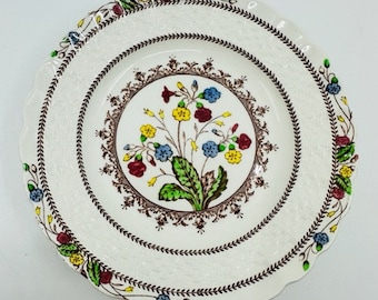 Op de afbeelding: Een wit keramisch dinerbord met een bruin, groen, geel, rood en blauw bloemenontwerp. Het bord heeft een gegolfde rand en een verhoogde rand.