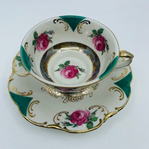 May include: A vintage teacup and saucer set with a white background, green and gold accents, and floral designs. The cup and saucer feature a delicate floral pattern with pink roses and green leaves. The rim of the cup and saucer is trimmed with a gold band.