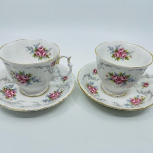 Könnte beinhalten: Zwei florale Teetassen und Untertassen mit einem zarten rosa-blauen Blumenmuster. Die Tassen und Untertassen haben einen goldenen Rand und einen weißen Hintergrund.