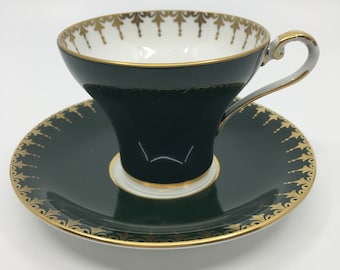 Può includere: Un set da tè composto da tazza e piattino verde e oro. La tazza e il piattino sono decorati con un bordo dorato e un delicato motivo floreale.