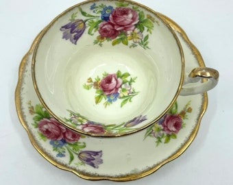 May include: A cream-colored porcelain teacup and saucer set featuring a floral pattern of pink roses, purple tulips, and small blue flowers. The rim of both the cup and the saucer is scalloped and finished with a metallic gold trim.