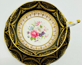 May include: A black and gold teacup and saucer set with a floral design. The saucer has a black rim with gold trim and a gold floral pattern. The teacup has a pink rim with gold trim and a floral design in the center.