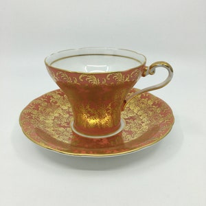May include: A porcelain teacup and saucer set featuring a coral-pink base colour with an intricate, reflective gold filigree pattern. The teacup has a scalloped rim, a white interior, and a curved handle with gold trim accents on the edges.
