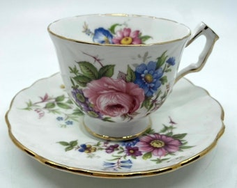 May include: A white teacup and saucer with a floral pattern in shades of pink, blue, and green. The cup has a gold rim and a delicate handle. The saucer has a scalloped edge and a gold rim.