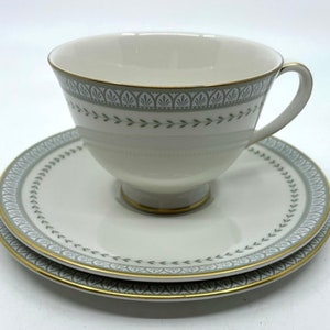 May include: A white teacup and saucer set with a delicate green and gold floral pattern. The cup has a rounded shape and a small handle. The saucer has a matching pattern and a slightly scalloped edge.