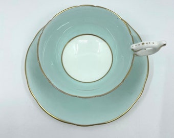 Può includere: Un set da tè composto da una tazza e un piattino di colore azzurro chiaro con un bordo dorato. La tazza da tè ha un interno bianco e un manico delicato.
