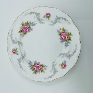 Könnte beinhalten: Weißer Porzellanteller mit gewelltem Rand und goldenem Rand. Der Teller hat ein Blumendesign mit rosa Rosen, blauen Akzenten und grauen Schnörkeln. Ein dekoratives Stück zum Servieren oder Ausstellen.