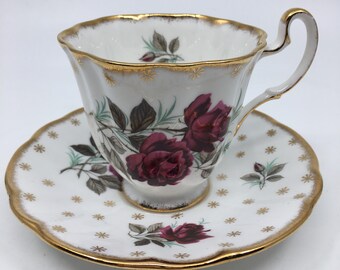Pode incluir: Um conjunto de chávena e pires branco com aro dourado e um desenho floral com rosas bordô. O pires tem um padrão de estrelas douradas.