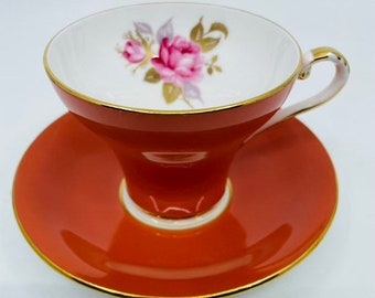 May include: A vintage teacup and saucer set with a floral design. The cup and saucer are both a vibrant orange color with a gold trim. The floral design features a pink rose with green leaves.