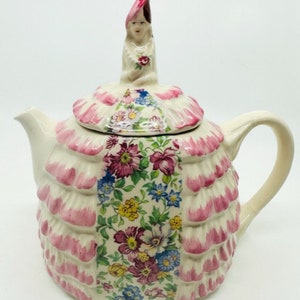 Puede incluir: Una tetera de porcelana vintage con un diseño floral y una figurita de una mujer con un vestido rosa en la tapa.