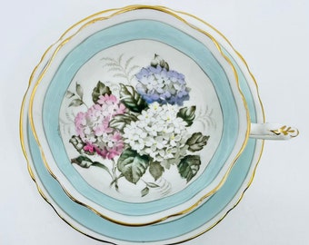 May include: A vintage teacup and saucer set with a delicate floral design. The cup and saucer are white with a light blue band and a gold trim. The floral design features hydrangeas in shades of pink, white, and purple.