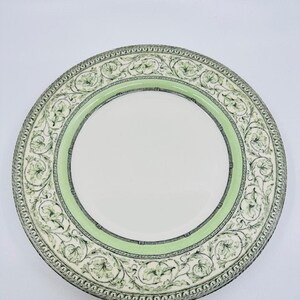 May include: A stack of white ceramic plates with a green and black floral design border. The plates have a light green inner border and a white center. The plates are stacked on a white surface.