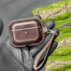 Könnte beinhalten: Ein braunes Lederetui mit einem genähten Rand und einer Metallschlaufe für einen Schlüsselanhänger. Das Etui hat eine Klappe, die mit einem Druckknopf geschlossen wird.