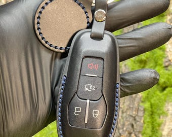 May include: Black leather key fob cover with blue stitching, attached to a key ring. The fob has buttons for lock, unlock, and other vehicle functions. A matching round leather tag is also attached.