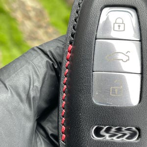 Op de afbeelding: Zwarte leren autosleutelhanger met rode stiksels. De sleutelhanger heeft drie knoppen met pictogrammen voor vergrendelen, ontgrendelen en het openen van de kofferbak. De autosleutel heeft een zilveren afwerking en het logo van het automerk.