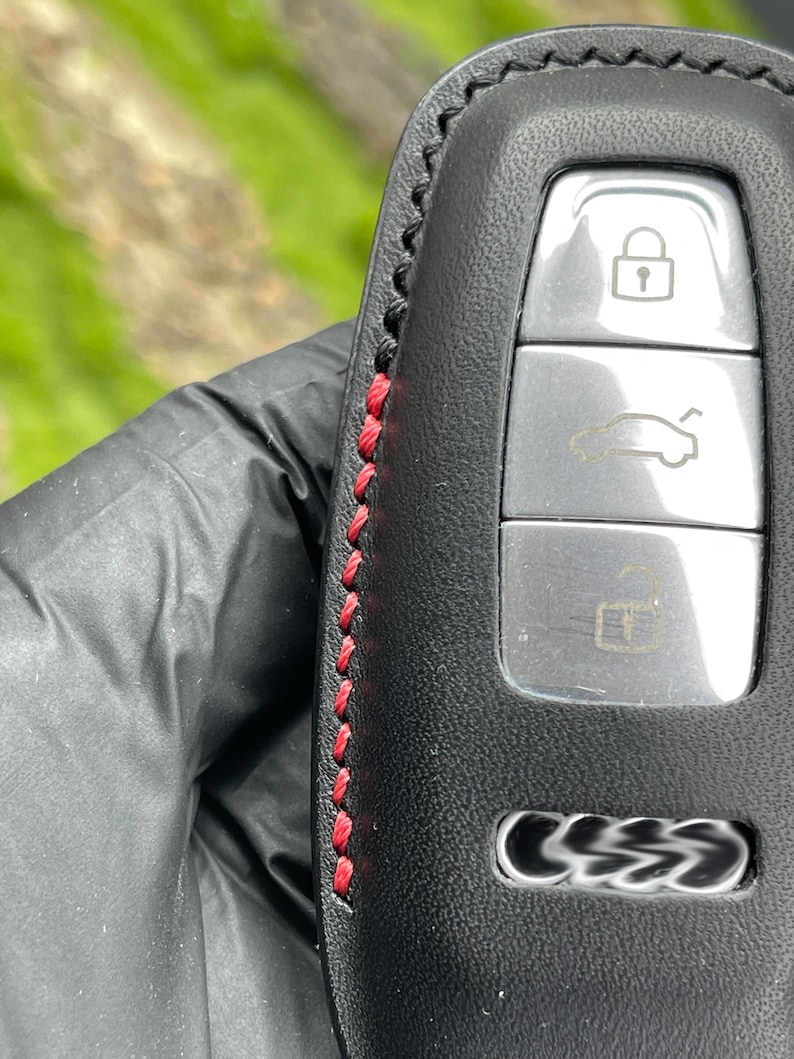 Op de afbeelding: Zwarte leren autosleutelhanger met rode stiksels. De sleutelhanger heeft drie knoppen met pictogrammen voor vergrendelen, ontgrendelen en het openen van de kofferbak. De autosleutel heeft een zilveren afwerking en het logo van het automerk.