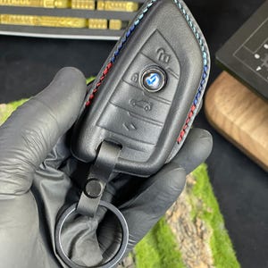 May include: Black leather key fob cover with red, white, and blue stitching. The fob has buttons for locking, unlocking, and other functions. A silver key ring is attached to the fob. The BMW logo is visible on the fob.