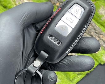 Könnte beinhalten: Schwarze Leder-Autoschlüssel-Hülle mit roten Ziernähten. Die Hülle hat silberne Knöpfe und einen schwarzen Schlüsselring. Der Schlüsselanhänger dient zum Schutz des Autoschlüssels.